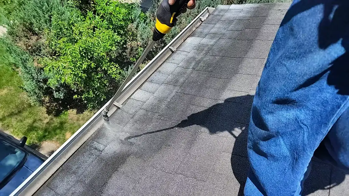 Spray wand working the shingles near a gutter line with greenery behind